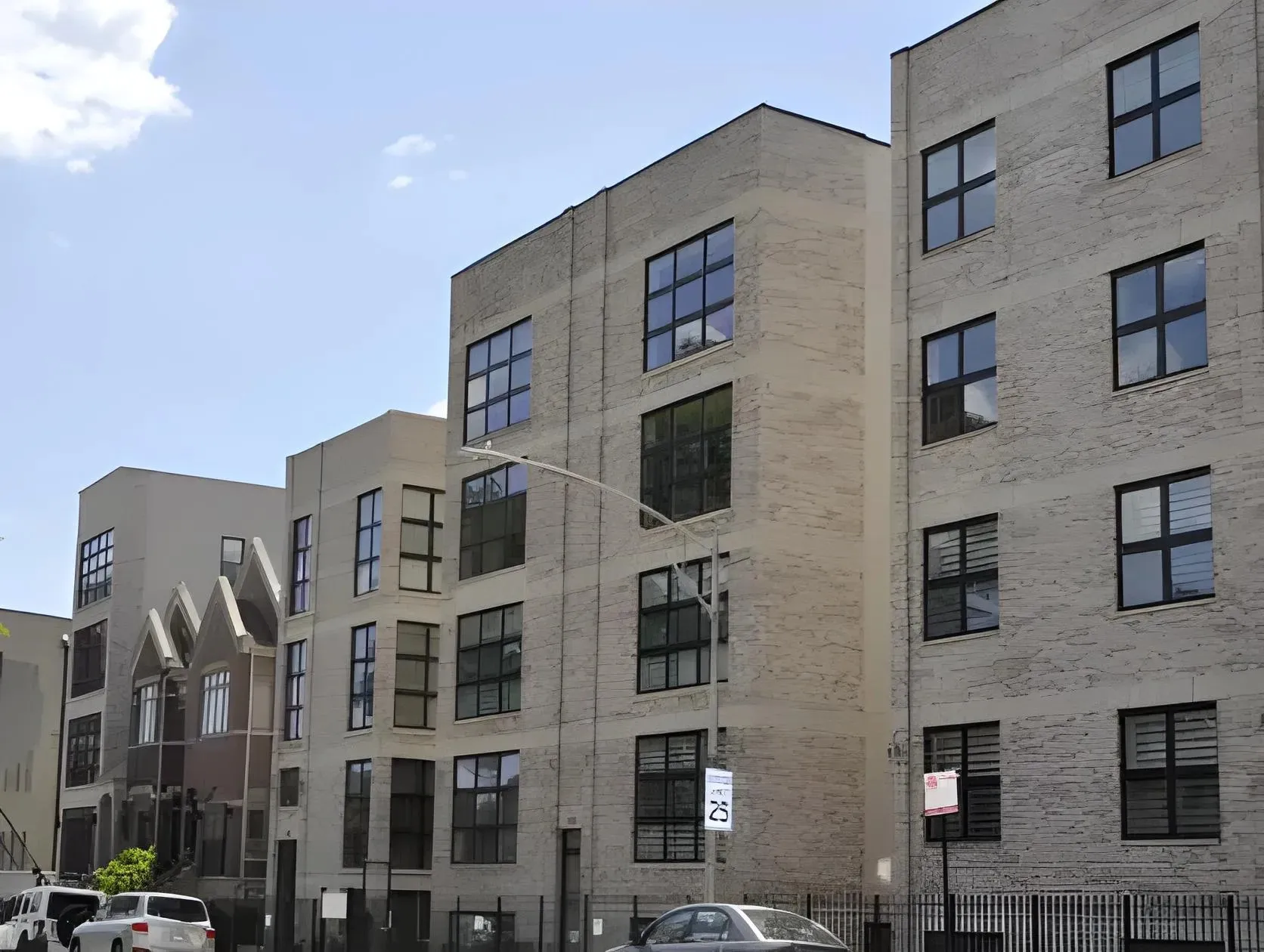 A street view of modern, multi-story apartment buildings with large windows. A few cars are parked along the sidewalk, and a street sign indicates a speed limit of 25 mph. The sky is clear with a few clouds.