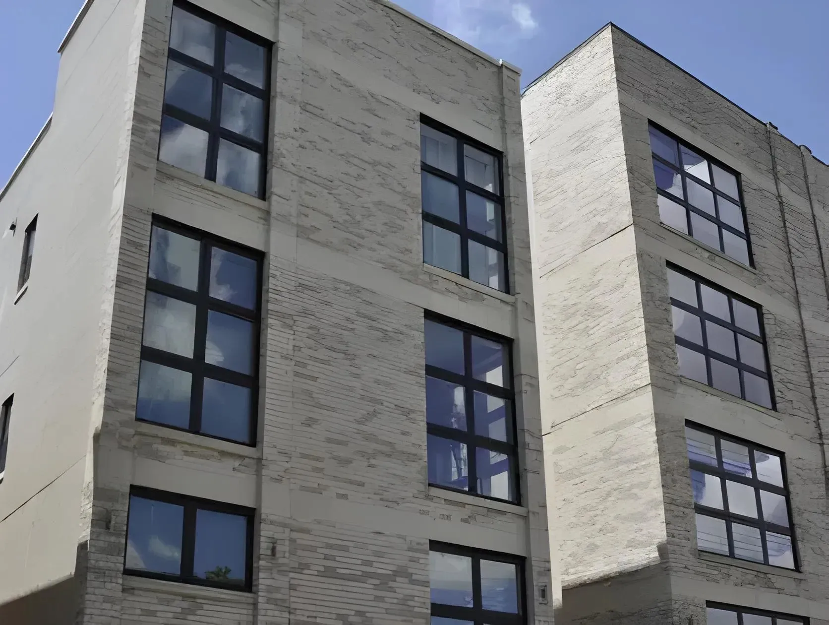 A close-up view of a modern, multi-story apartment building with large, grid-patterned windows. The exterior features a combination of light-colored bricks and smooth surfaces. A clear, blue sky with a few clouds is visible in the background.