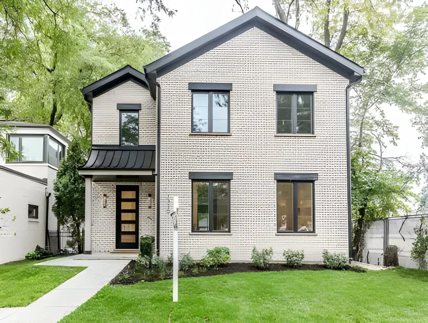 A modern two-story house with a light brick facade and dark window frames. The house features a sleek black door and a small entrance canopy. It is surrounded by a well-maintained lawn and trees, with a concrete pathway leading to the entrance.