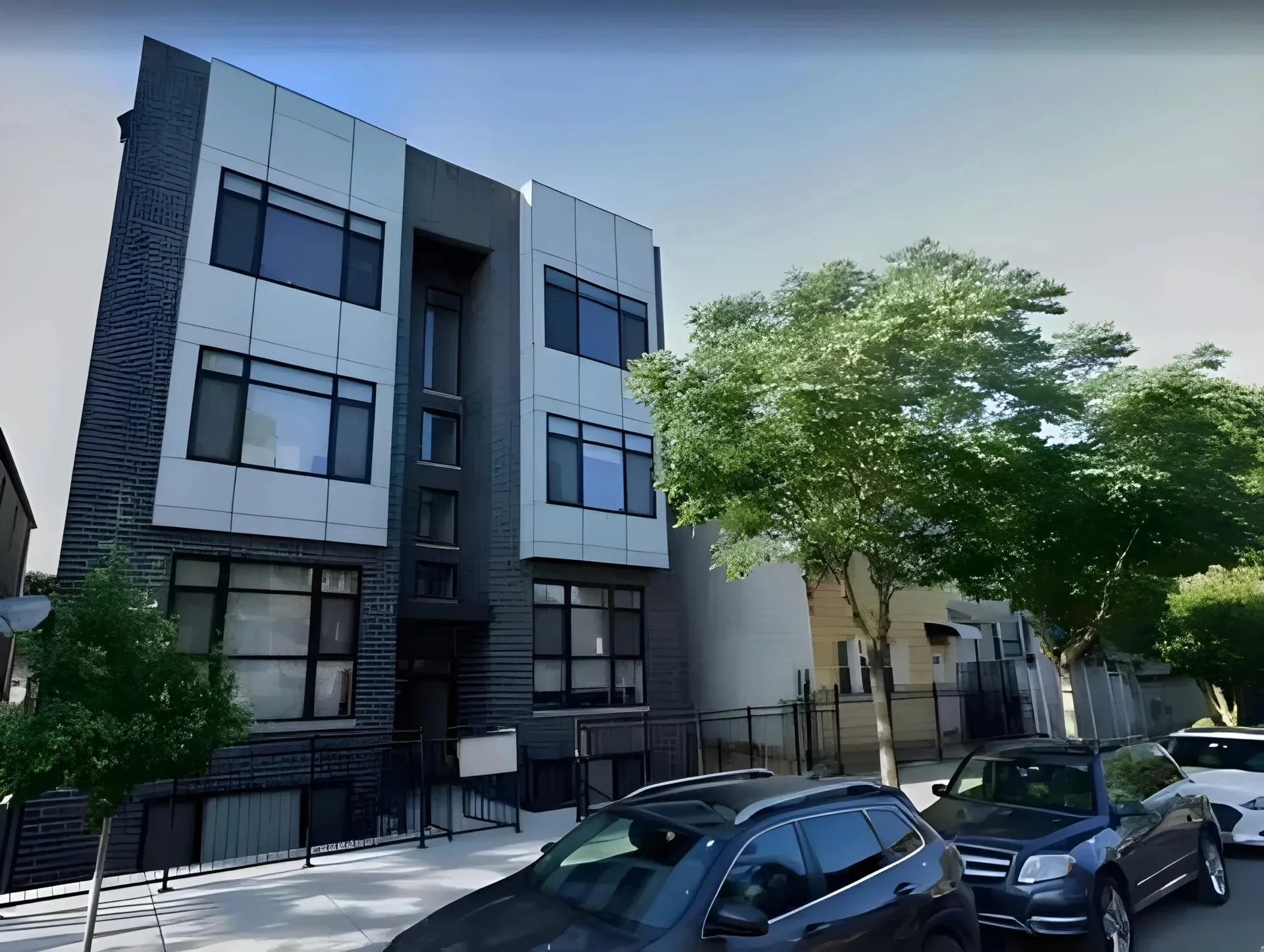 A modern three-story building with large windows and a combination of white and dark exterior panels. There are two trees in front, parked cars along the street, and a sidewalk with a low metal fence lining the building's perimeter.