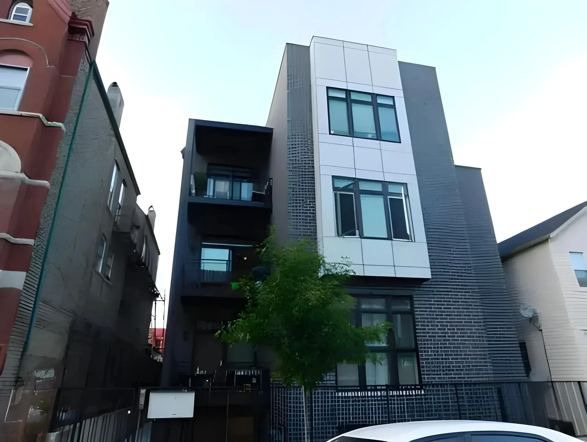 A modern, three-story apartment building with a mix of white, dark gray, and brick exterior. The structure features several large windows, a balcony on the left, and a small tree in front. Adjacent buildings partially visible on either side.