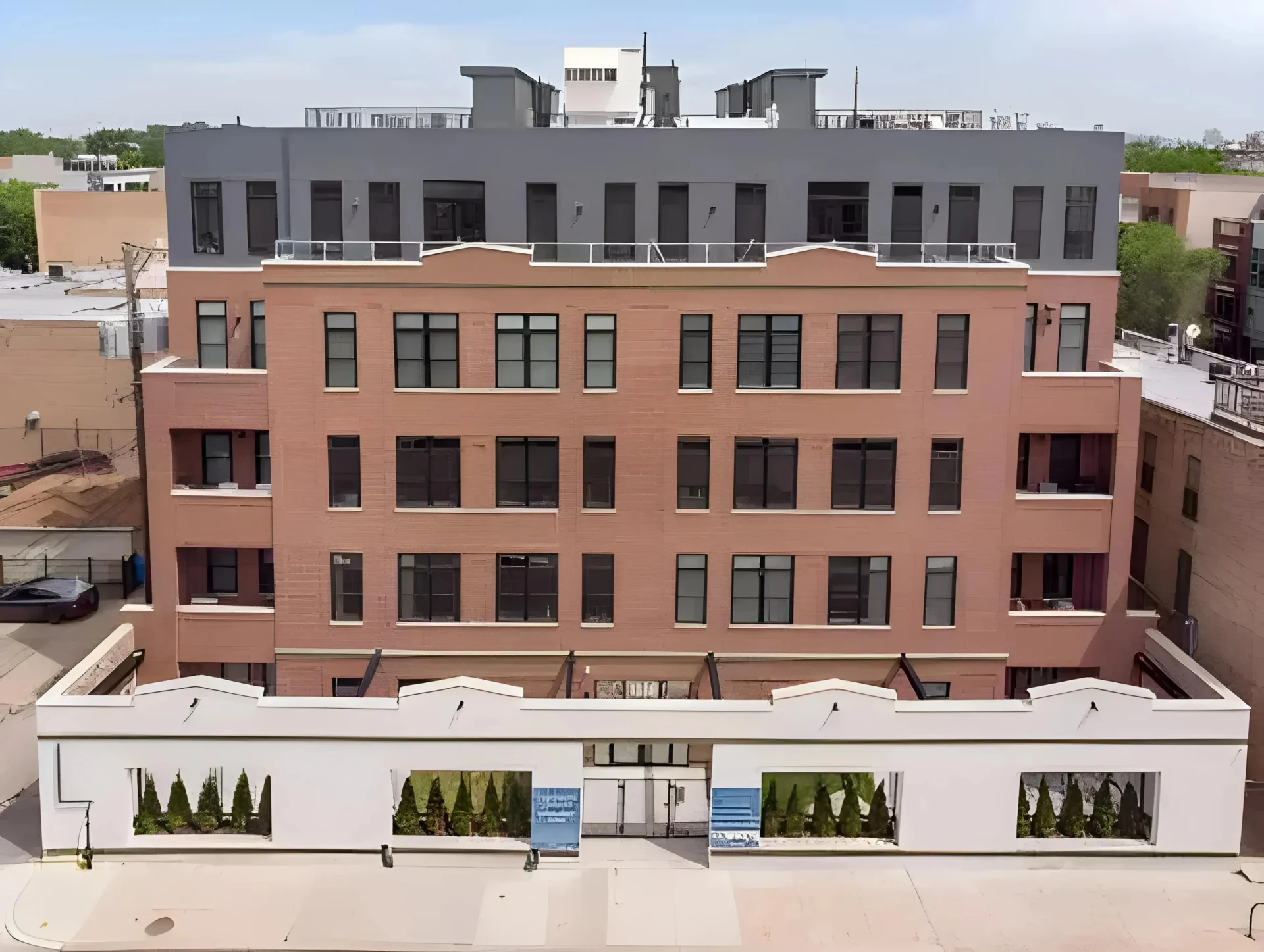A multi-story brick and stucco building with large windows and a flat roof, possibly apartments or offices. The lower level has a gated entrance with green shrubs lining the exterior. The building is situated in an urban area.