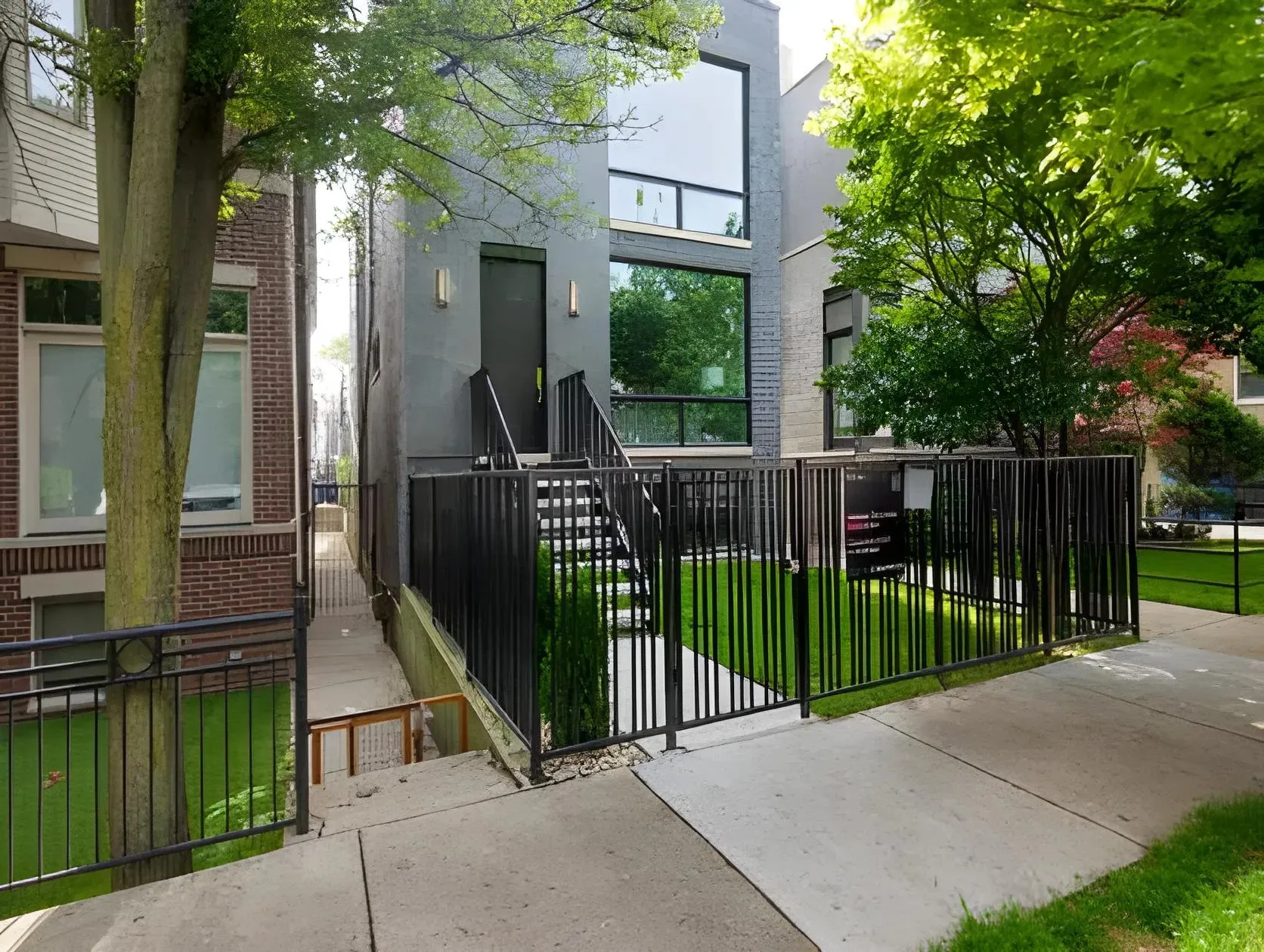 A modern, two-story gray house with large windows is situated between two other homes. A black metal fence surrounds the front yard, which has green grass and a tree. Concrete steps lead up to the entrance, accessible via a gated walkway.