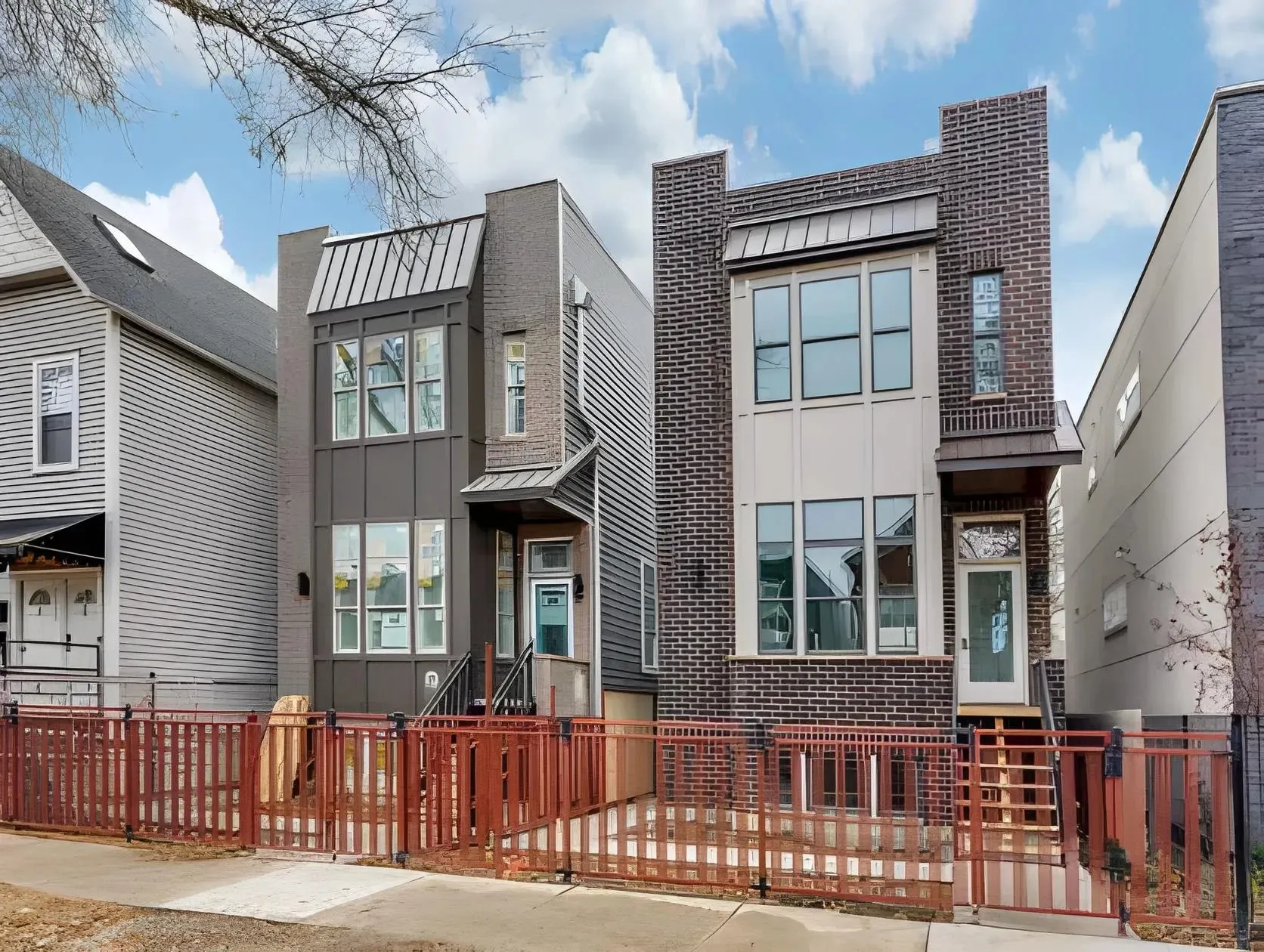A modern residential street view featuring two adjacent multi-story houses with large windows. The home on the left has a combination of gray and brick exterior while the one on the right features red brickwork and beige accents. A red fence runs in front.