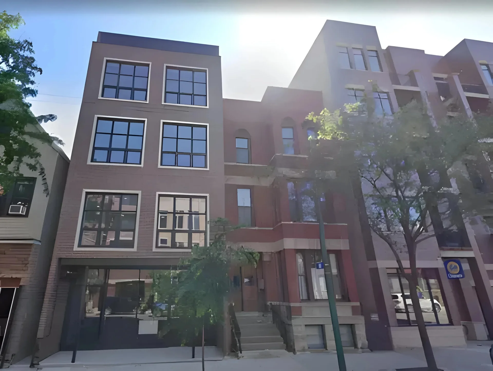 Four-story buildings with large windows stand side by side on a sunny day. The structure on the right has red brickwork and a staircase leading to the entrance, while the building on the left has a glass storefront and a modern facade. Trees line the sidewalk.