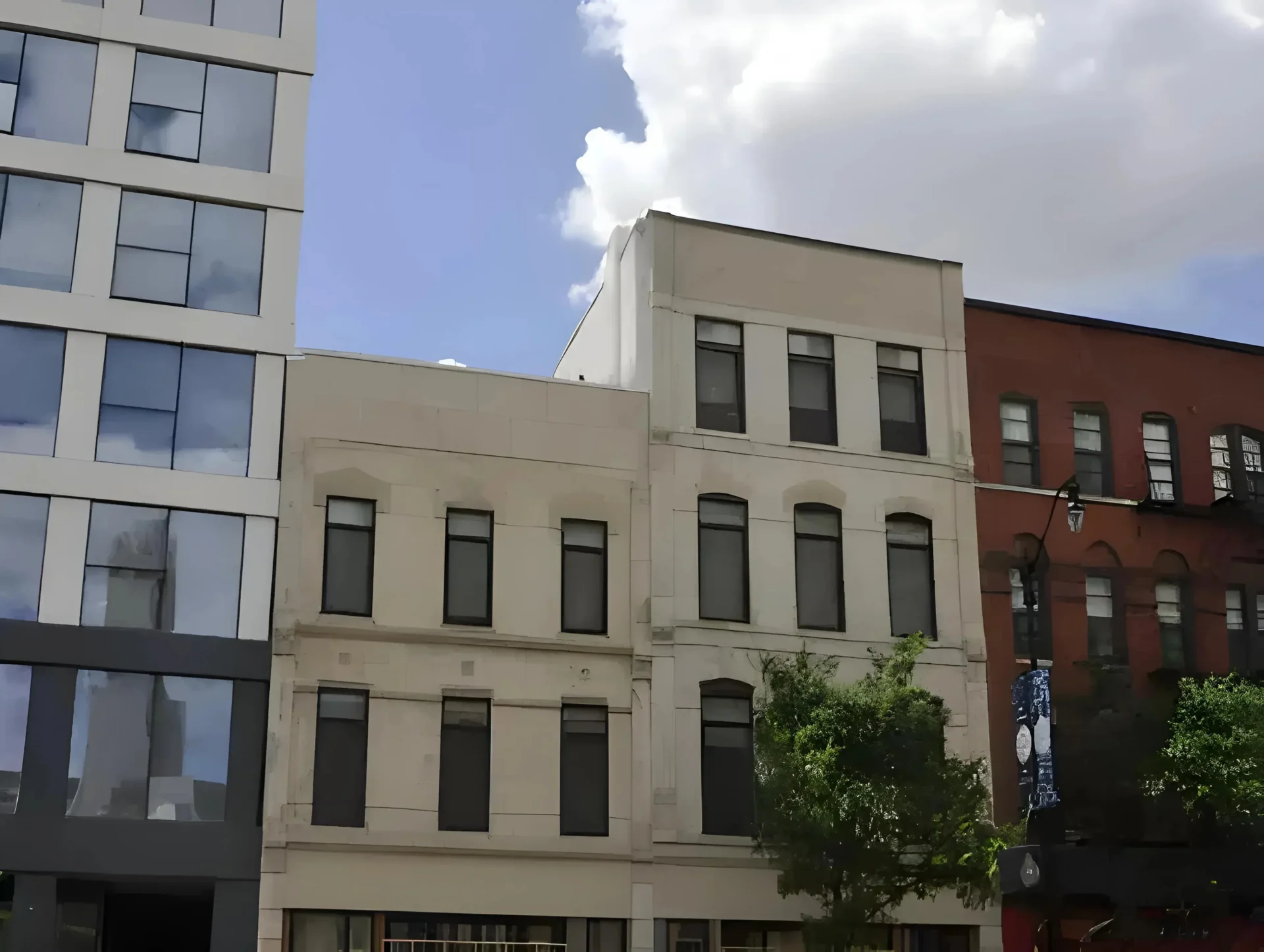 A street view of a row of buildings, including modern and older structures. The central building is a beige, three-story structure with multiple windows. A red-brick building is partially visible on the right, and a tree is in front of the buildings. Blue sky with clouds above.