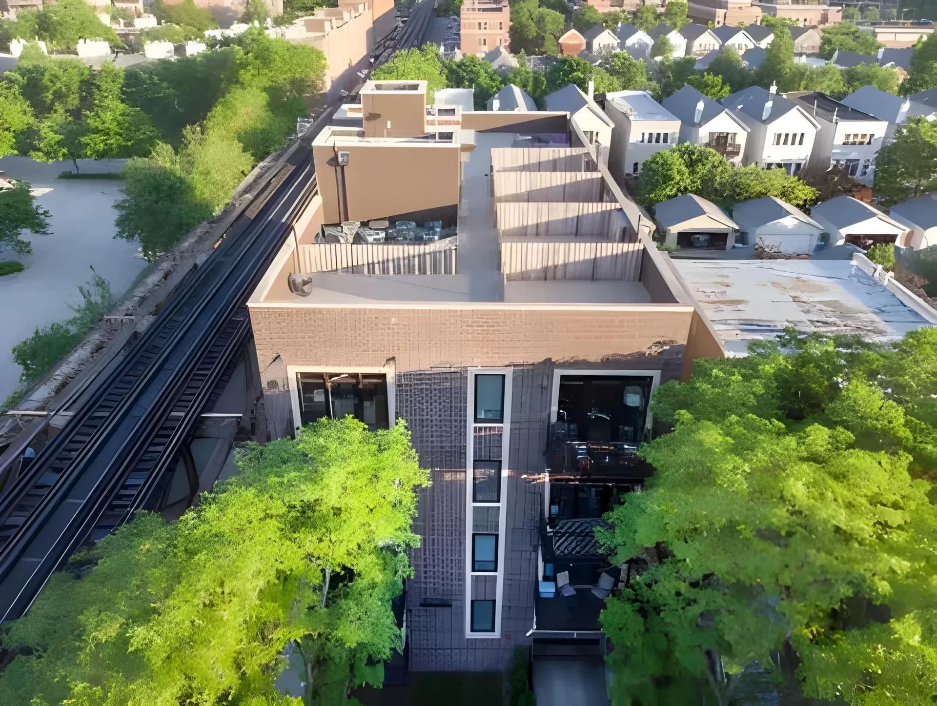 An aerial view of a residential area featuring a modern, multi-story building adjacent to a railway track. The building has balconies and is partially surrounded by lush, green trees. In the background, there are other homes with white roofs, and the area is densely populated with greenery.