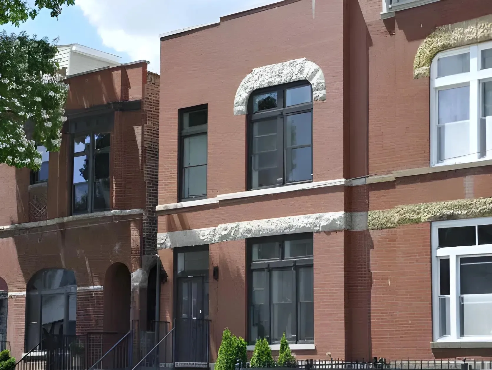 A brick townhouse with large windows and stone accents. The building features a modern design with clean lines. To the left, a neighboring brick building has an arched entryway and similar windows. A small black fence surrounds the front yard.