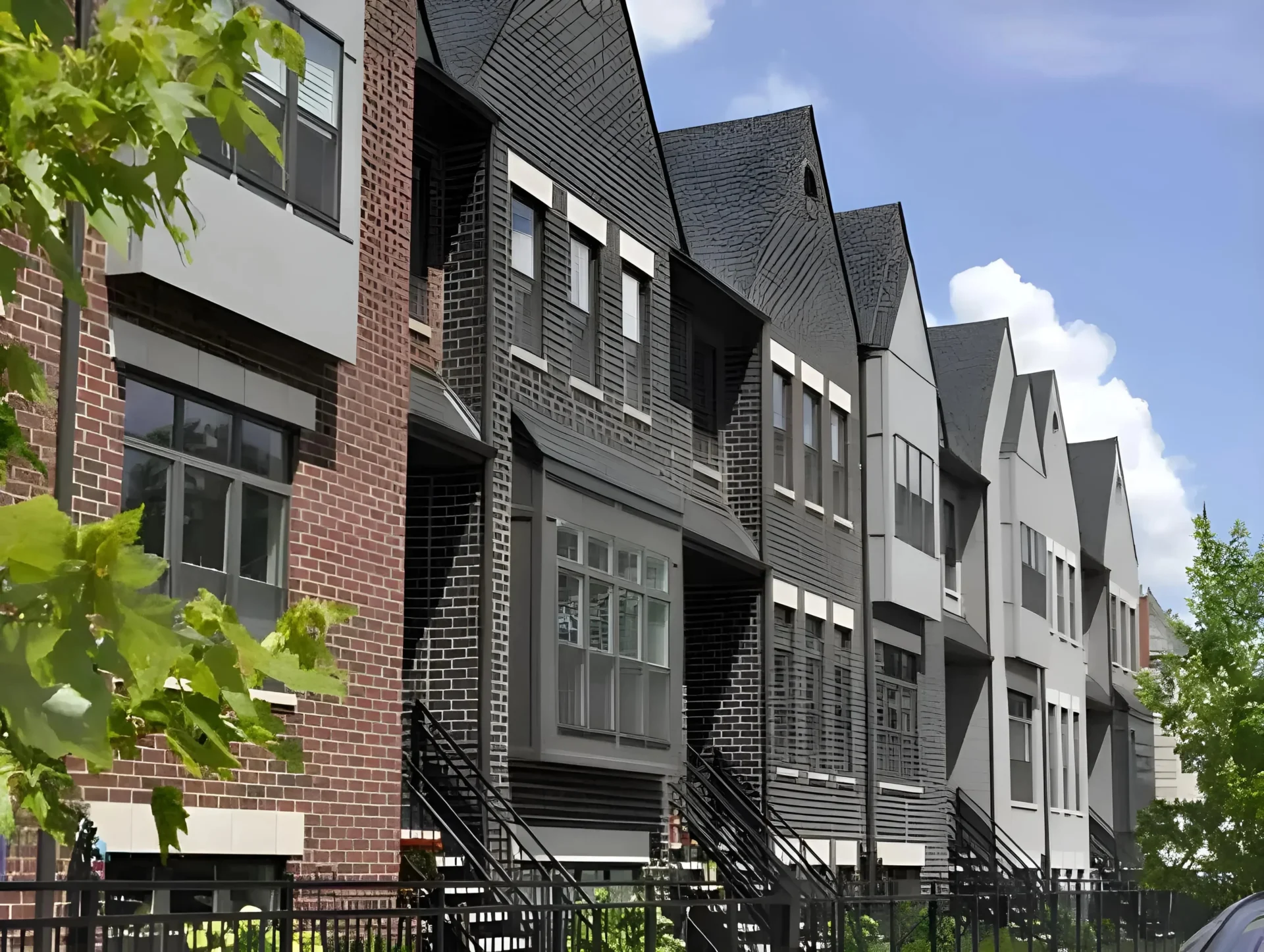A row of modern townhouses with a brick and gray facade, featuring large windows and black metal stair railings. The homes are adjacent to leafy green trees and shrubs, with a clear blue sky in the background.
