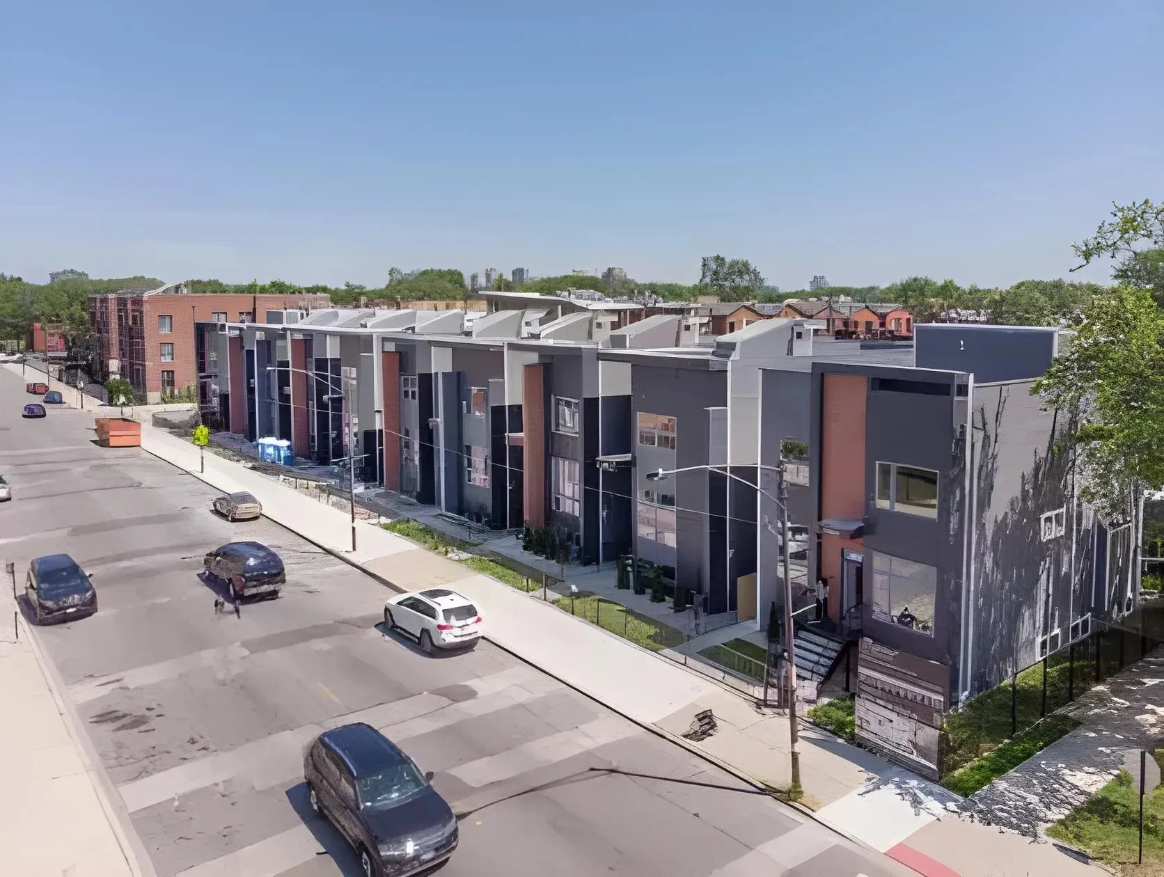A street view of a modern suburban neighborhood with a row of contemporary townhouses on the right. Several cars are parked along the curb and driving on the road. The townhouses feature a mix of grey and red brick exteriors with white accents.