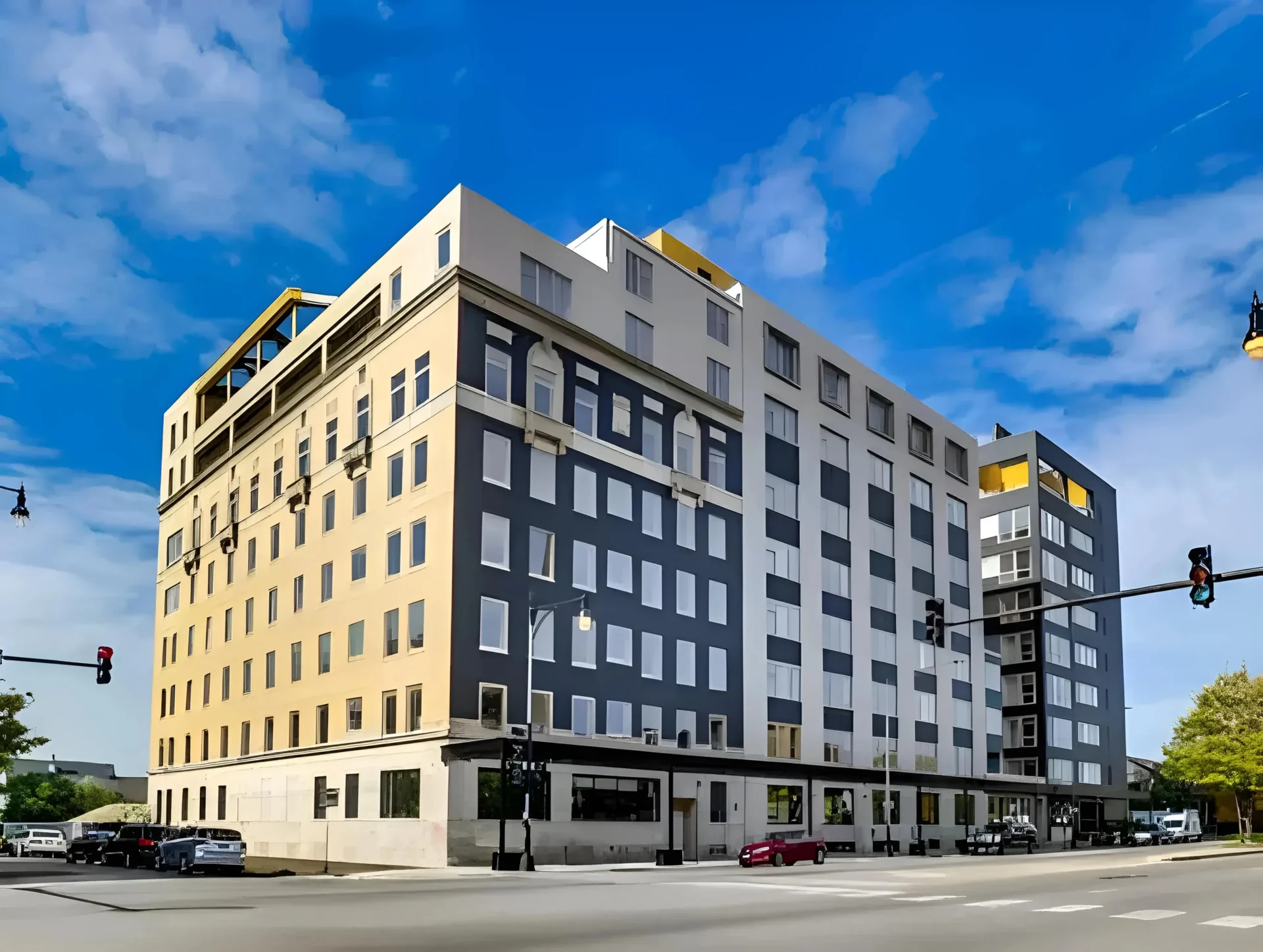A modern multi-story building with a beige and gray facade stands at a street intersection. The building has numerous windows, a few small balconies, and architectural accents in yellow. Traffic signals and street lamps are visible, with a bright blue sky overhead.