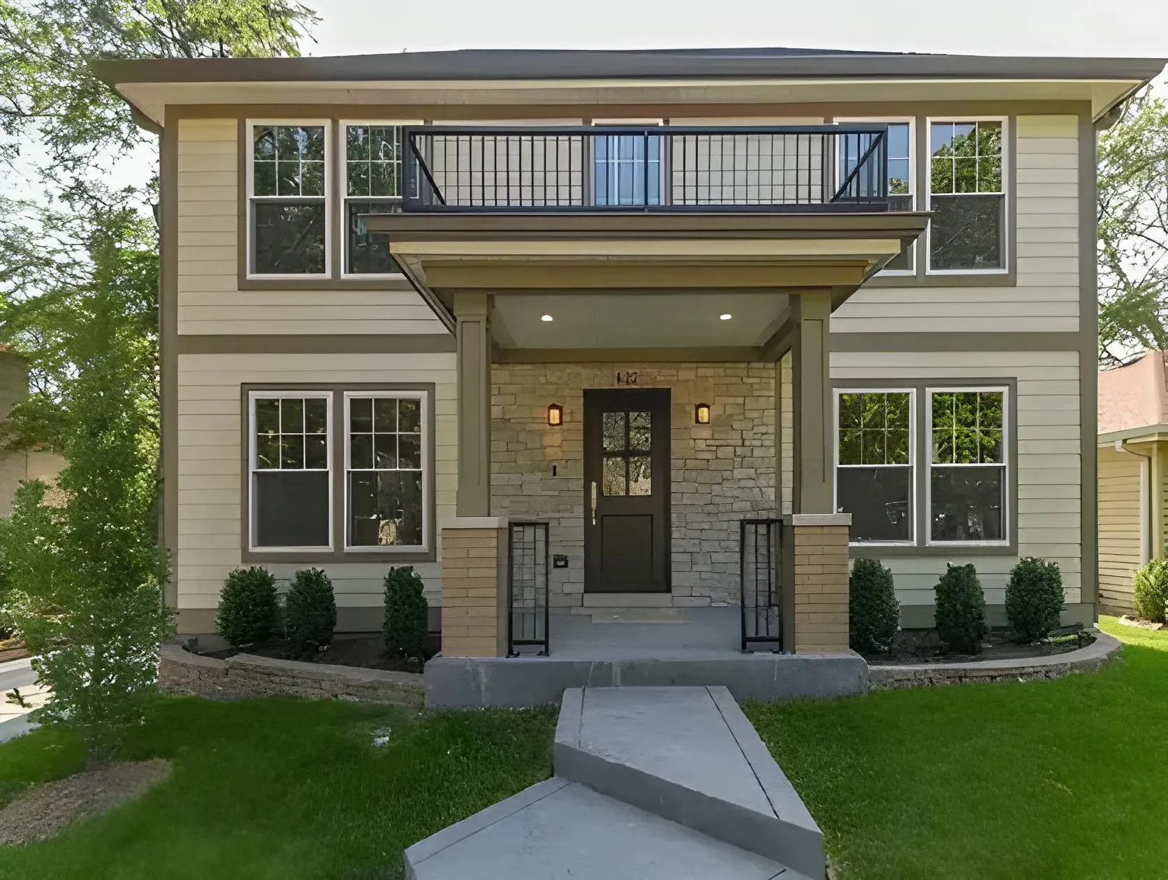 A two-story house with a modern design features a front entrance with a wooden door, stone and brick accents, and a small balcony above. The house includes large windows on both floors, a concrete walkway leading to the front porch, and well-manicured landscaping.