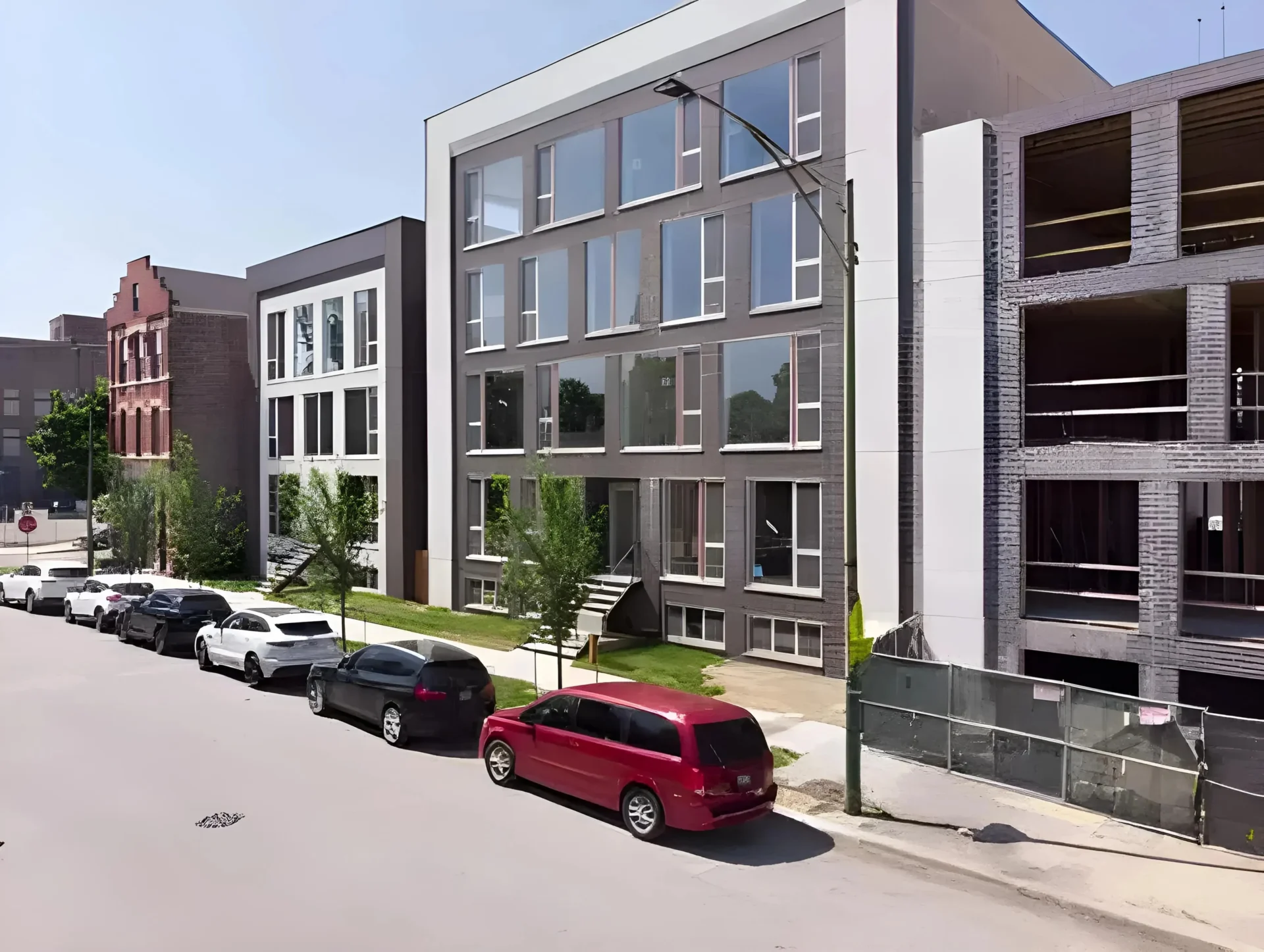 A row of modern, multi-story apartment buildings with large windows stands along a street. A brick building is visible in the background. Several cars are parked along the curb, including a red van, white car, and black SUV. Construction is ongoing nearby.