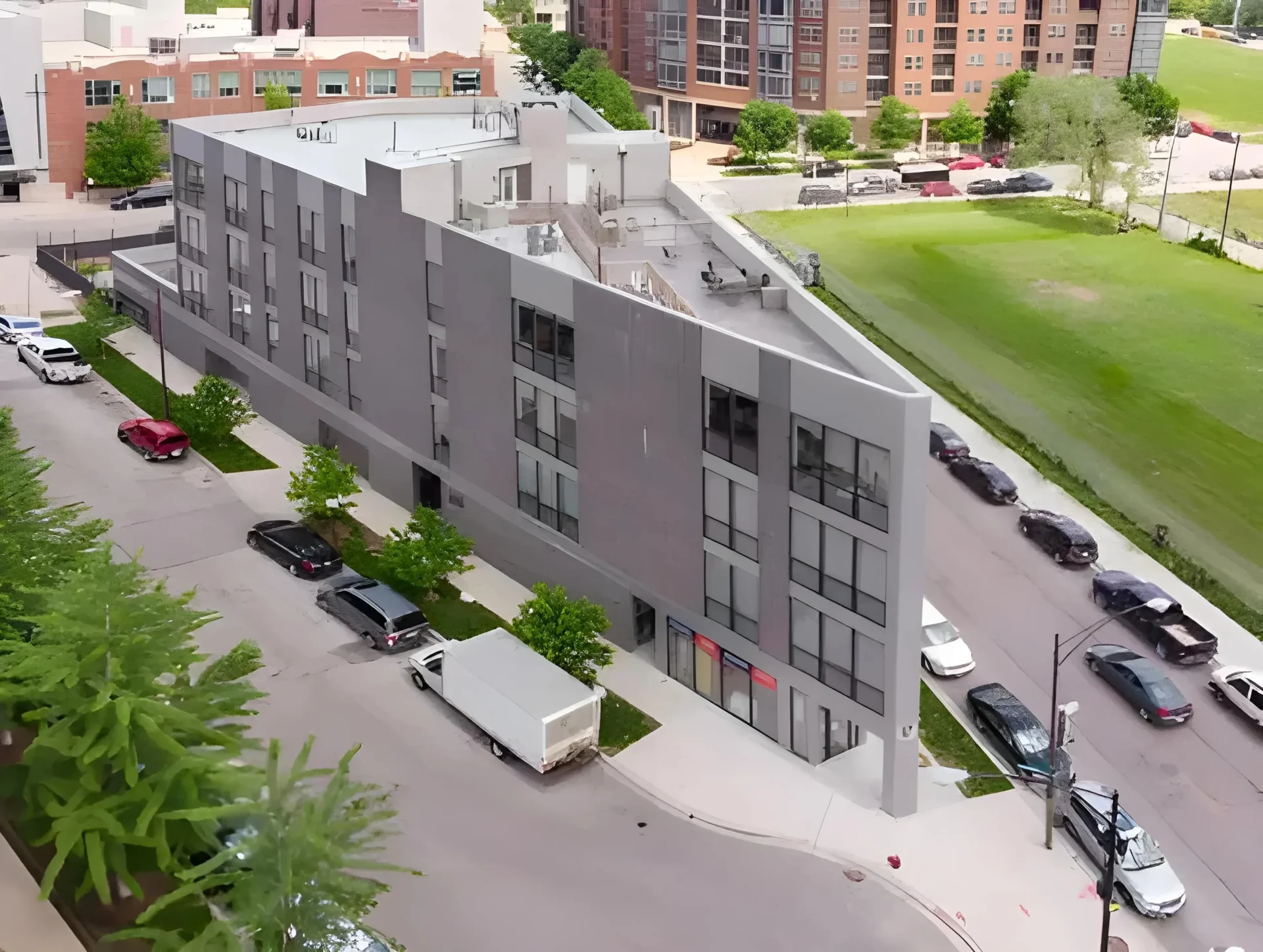 Aerial view of a modern, angular, four-story gray building situated at a corner. Several parked cars and a white moving truck are visible along the adjacent streets. The area is surrounded by green spaces, trees, and other buildings in the background.