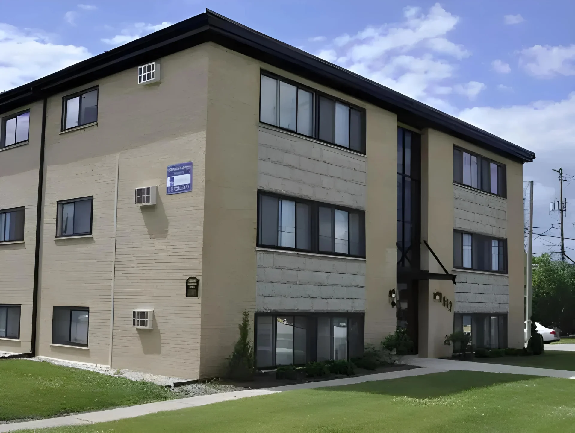 A three-story beige apartment building under a blue sky. The building has multiple windows with some air conditioning units installed. A sidewalk leads to the entrance, and a patch of green grass surrounds the property. A "For Rent" sign is visible on the side.