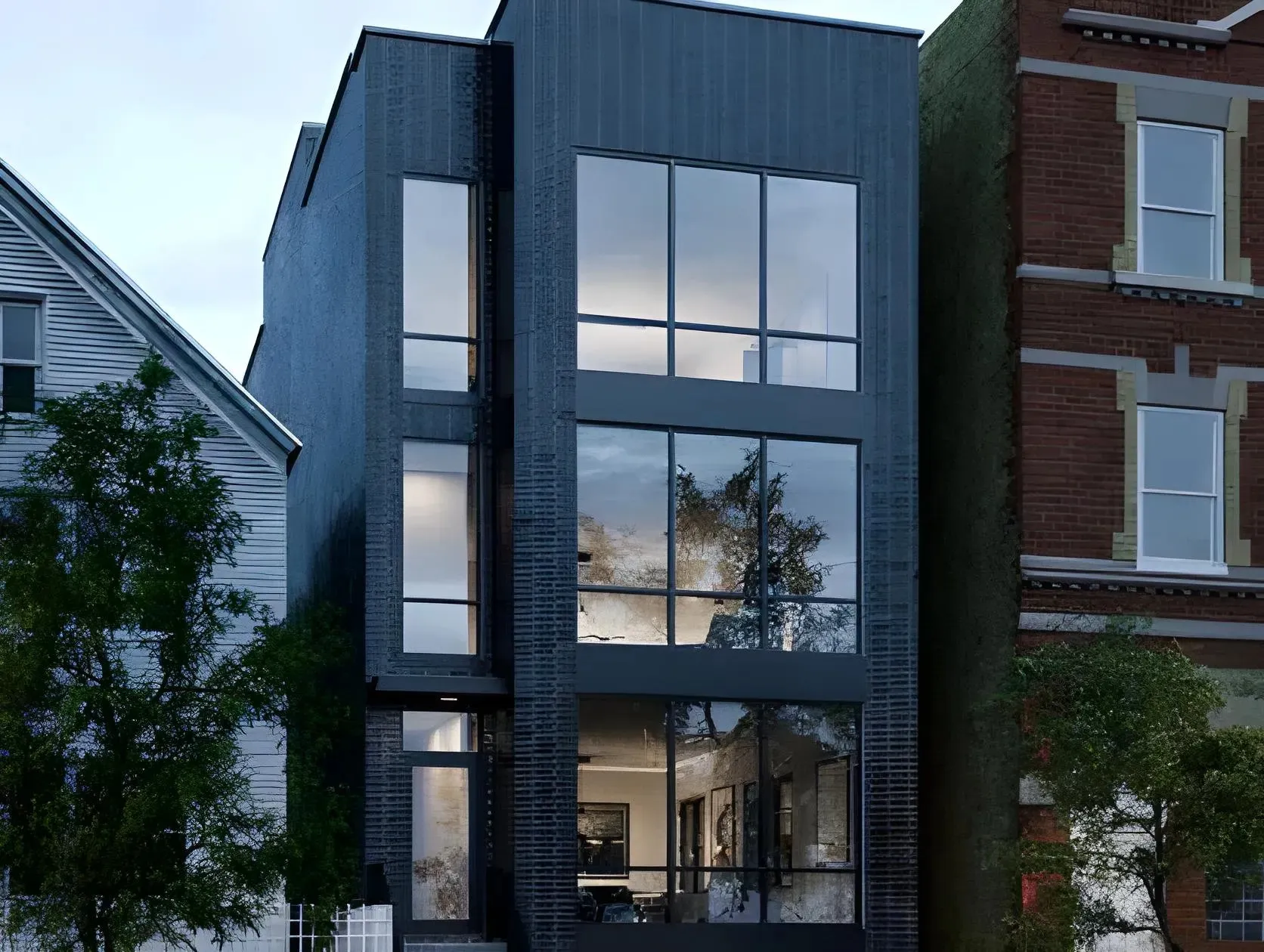 A modern, multi-story building featuring large glass windows and a sleek, dark exterior. The building is situated between two other structures, with trees visible in the front. The interior appears well-lit and contemporary.