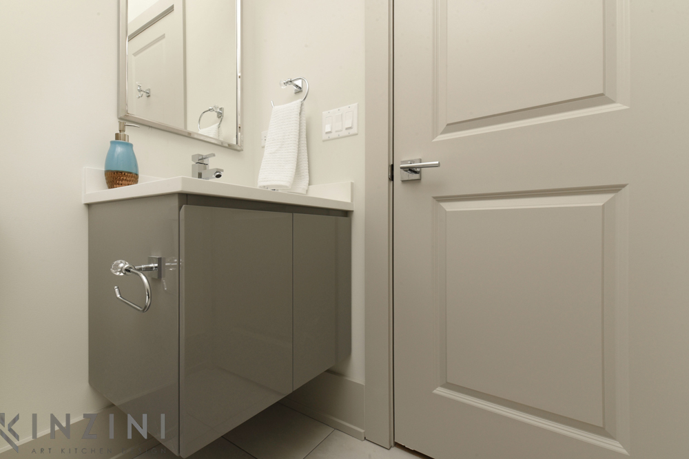 Modern bathroom corner with a sleek, gray vanity and a white countertop. The sink has a soap dispenser and a towel is neatly hung on the wall. The room features a large mirror and a closed door, all in neutral tones.